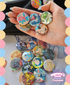 Hand holding colorful buttons with various designs, including pokemon characters, against a multicolored circular border.